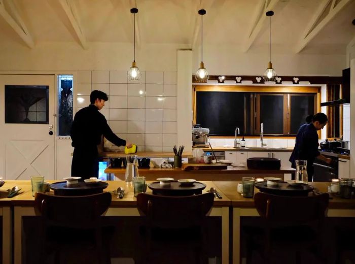 In an otherwise vacant kitchen, two staff members prepare dishes, visible beyond a tasting counter set for guests.