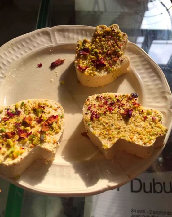 An overhead view of a plate displaying three pieces of halva molded into hamsas (hands), adorned with rose petals and pistachio bits.