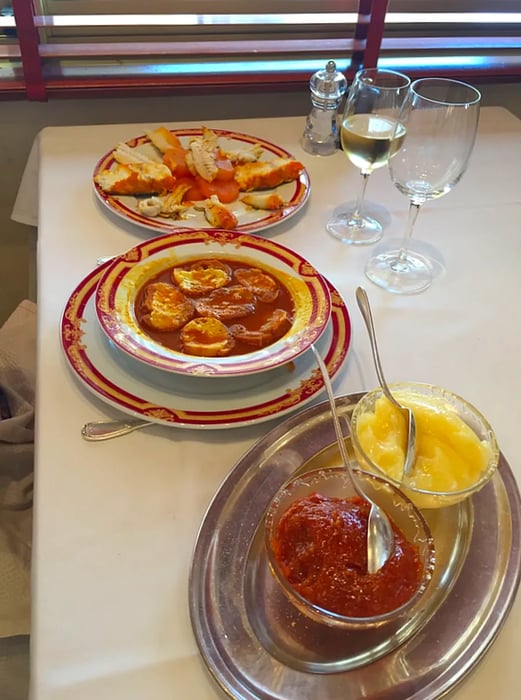 A bowl of bouillabaisse is placed at the center of a table, flanked by a platter of cooked fish and shellfish, along with a serving dish containing two bowls of condiments, all set beside wine glasses in front of a window with partially drawn blinds allowing streams of light to filter in.