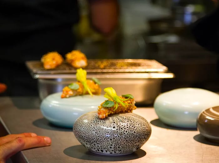 Two small dishes are beautifully presented on round stone-like bases resting on a steel prep station, with a chef's hands nearby.