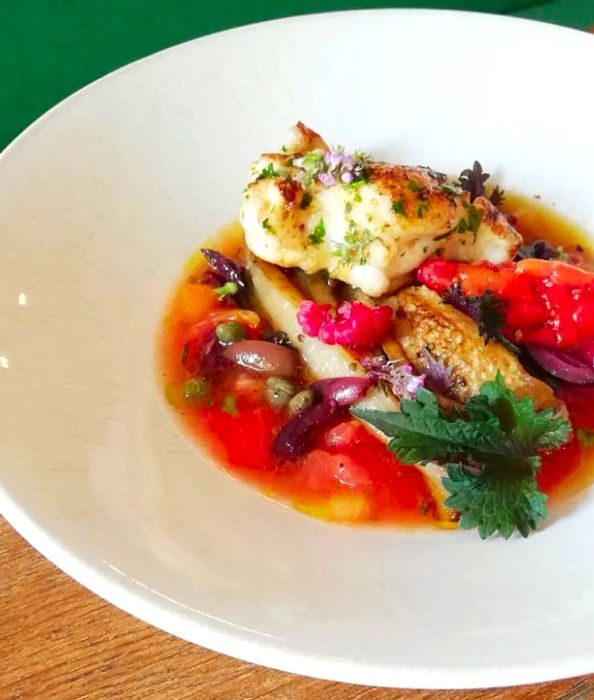 A dish features cooked fish nestled in a flavorful broth, complemented by eggplant, peppers, and raspberries among other vegetables.