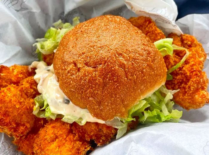 Aerial view of a sandwich overflowing with fried chicken protruding from both sides of a round bun, accompanied by mayo and lettuce, served in paper.