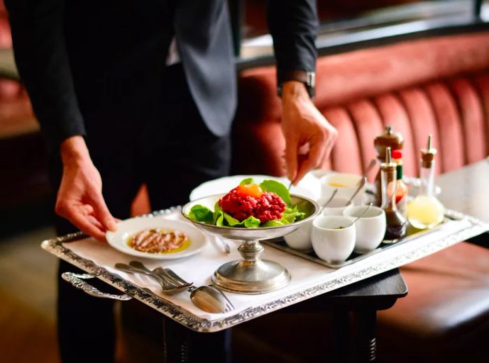 A server presents steak tartare on a trolley, accompanied by various sauces and condiments.