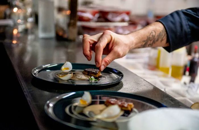 A hidden chef artfully arranges a final ingredient on one of several plates lined up on a bar, with the kitchen prep areas softly blurred in the background.