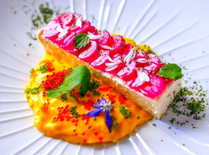 A long, rectangular piece of fish adorned with vibrant vegetable medallions, all resting in a luminous saffron cream sprinkled with various garnishes