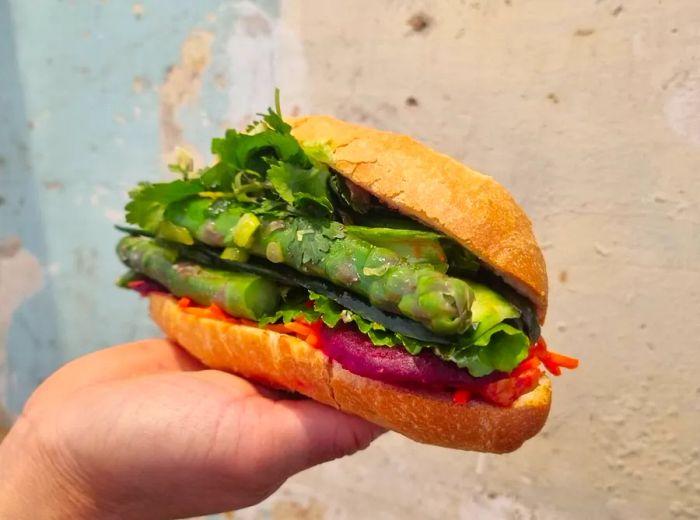 A hand presents a banh mi brimming with asparagus, mustard greens, and other vegetables against a concrete wall.