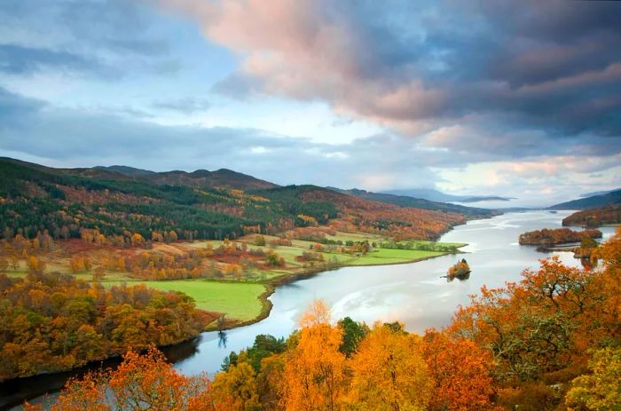 A Scottish loch near Pitlochry is surrounded by rolling farmlands and hills adorned with a mix of green and autumn-hued leaves.