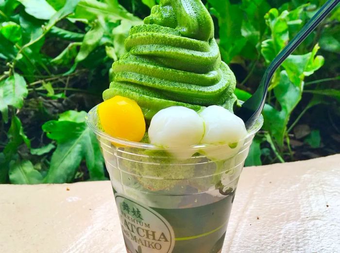 A plastic cup brimming with mochi, cake, and various fillings, topped with a generous swirl of matcha soft serve and a plastic spoon poking out.