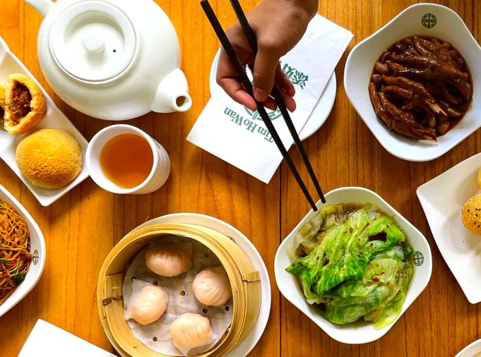 From above, a diner reaches for a dish of leafy greens, surrounded by choices of noodles, dumplings in a steamer, and more.