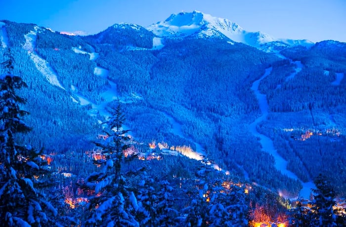 A breathtaking view of the mountains at dusk, with ski slopes visible above a town that’s illuminated at night.