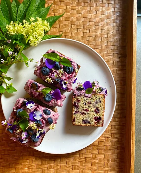 Aerial view of a loaf cake adorned with vibrant purple icing, whole blueberries, and sprigs of herbs.