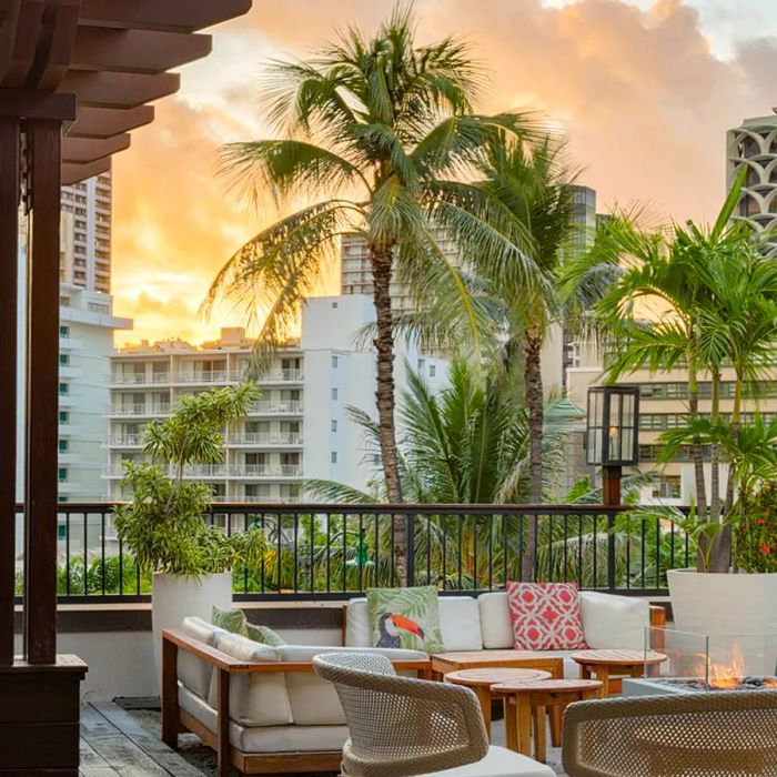 An outdoor lounge area adorned with cushioned seating and four-top tables, set against a backdrop of a sunset sky with palm trees and towering buildings.