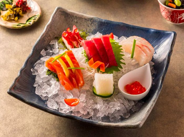 A square slate bowl filled with sliced sashimi on ice, accompanied by rice, pickles, and tea.