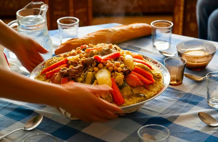 A traditional Moroccan homemade couscous dish presented on a blue checkered tablecloth, served by lively hands with glasses, spoons, and Laban milk.