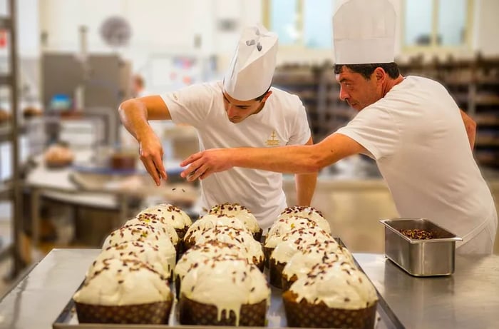 Two bakers dressed in white shirts and hats sprinkle toppings on panettoni.