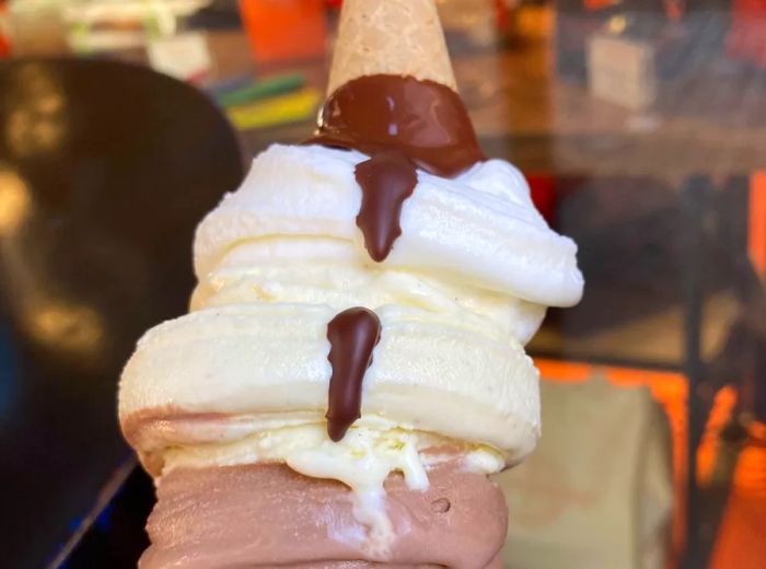 A cone layered with multiple flavors of ice cream, finished with a drizzle of chocolate and crowned with an upturned mini cone.