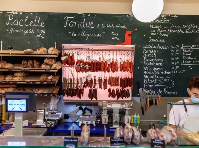 Cured meats hang beside shelves filled with bread, as an employee attends to customers behind the counter.