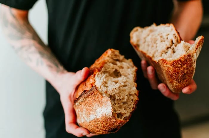 A baker displays two halves of a torn sourdough loaf towards the camera.