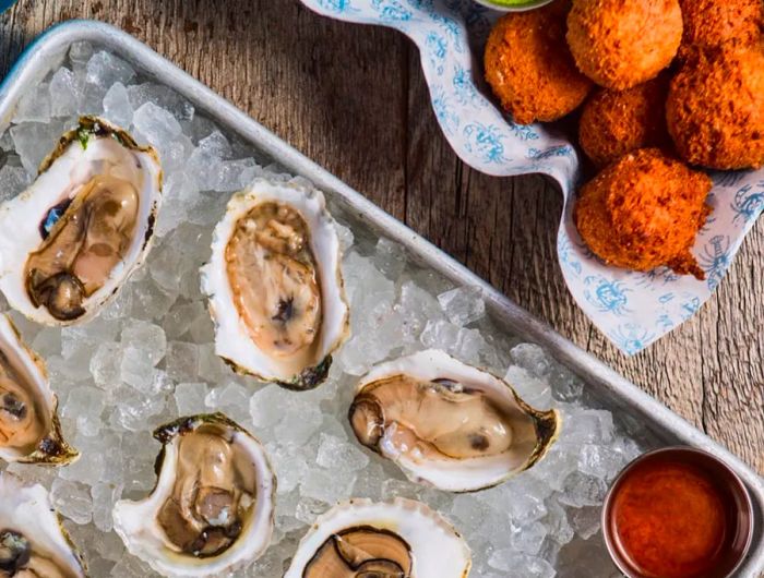 An aerial view of oysters resting on ice, accompanied by sauces and a basket of hush puppies.