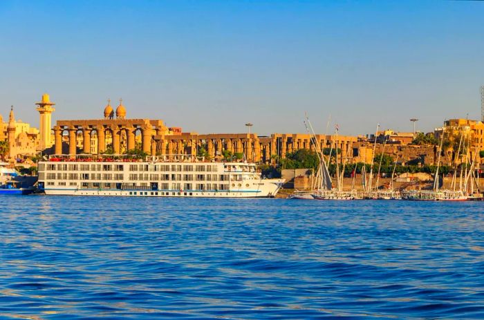 A large cruise ship alongside numerous small sailboats is docked along a river, with a majestic ancient temple complex in the background.