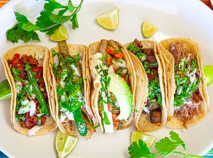 A platter of tacos, some filled with meat and others with vegetables, peppers, and herbs, served with lime wedges