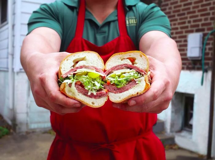 A chef in an apron stands in an alley, presenting a halved sandwich showcasing salami, lettuce, and cheese.