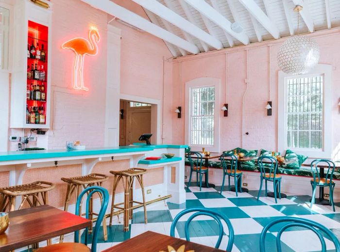 The interior of a restaurant features a checkered floor, wooden tables with curved backs, a bar lined with wooden stools, and a neon flamingo amid bottles, all illuminated by sunlight streaming through the windows.