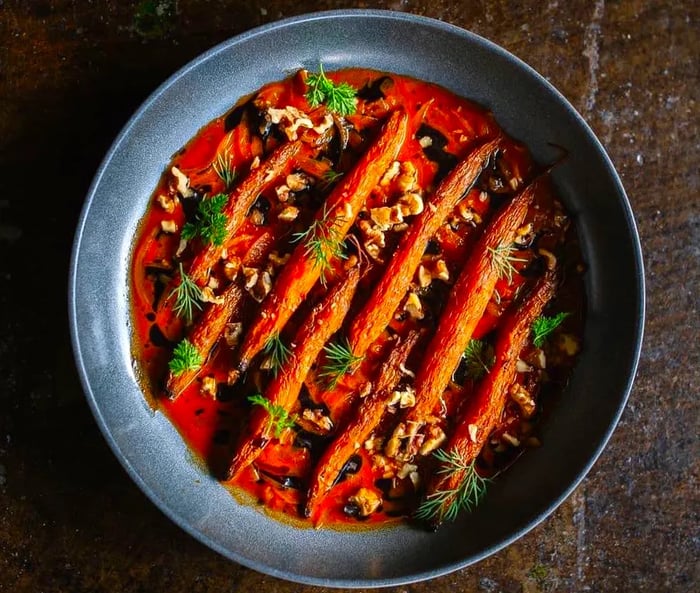 A slate-colored plate showcases long carrots in sauce, garnished with herbs and nuts, set against a textured stone background.