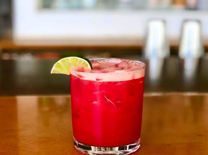 A vibrant red cocktail in a lowball glass, adorned with a lime wedge, sits on a rustic wooden bar.