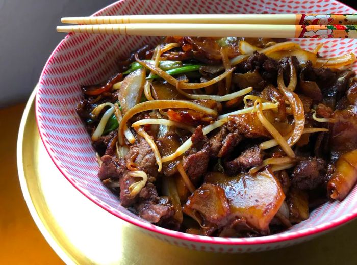 A decorative bowl of chow fun featuring slices of beef and fresh vegetables, beautifully presented on a charger