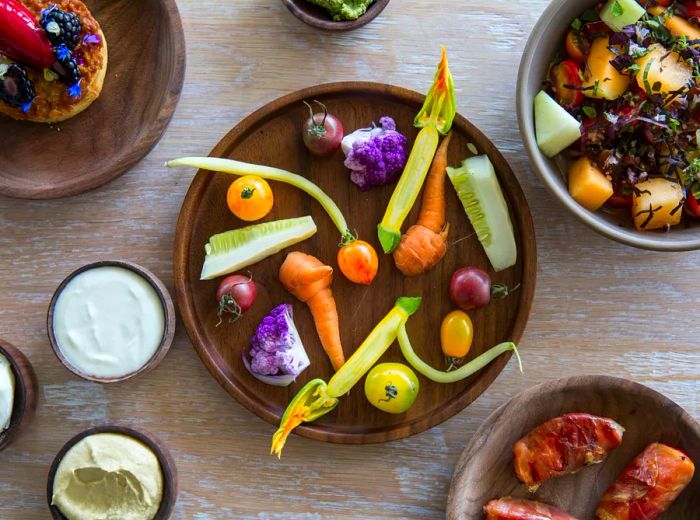 An overhead view of a platter featuring crudités with dips, a fruit tart, a tomato salad, and bacon-wrapped dates.