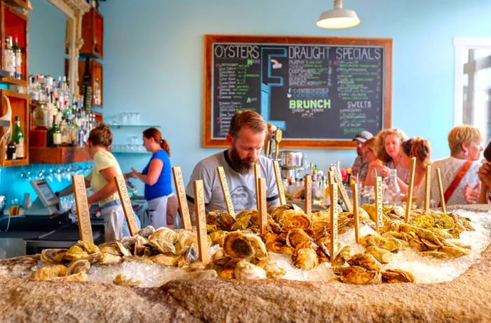 In the foreground, a raw bar brimming with oysters is visible, while diners enjoy their meals in a casual restaurant setting in the background.