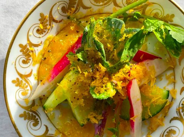 An overhead view of a salad featuring sliced cucumbers, radishes, and greens drizzled with a thick orange sauce, elegantly arranged on a decorative plate.