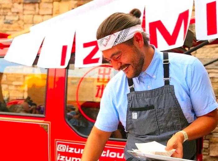 A chef wearing a headband stands next to a small red firetruck, with a large banner that reads Izumi.