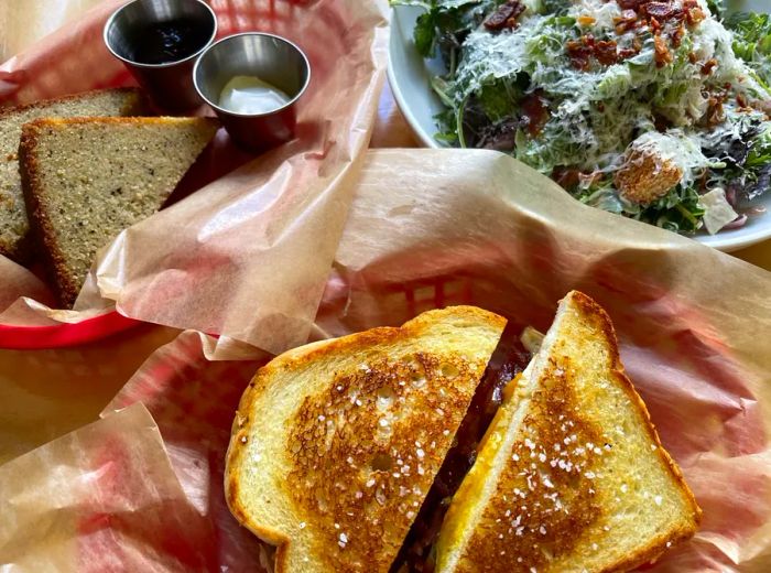 An overhead view of a sliced grilled cheese served in a paper-lined basket, alongside a salad and another bread-based dish.