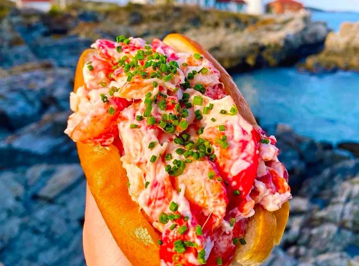 A hand holds a generously packed lobster roll, topped with mayo and chives, set against a backdrop of rocky coastline and a distant lighthouse.