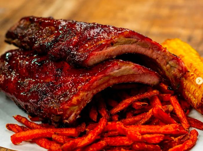 A platter featuring two racks of glistening ribs resting on butcher paper alongside sweet potato fries and a corn cob.