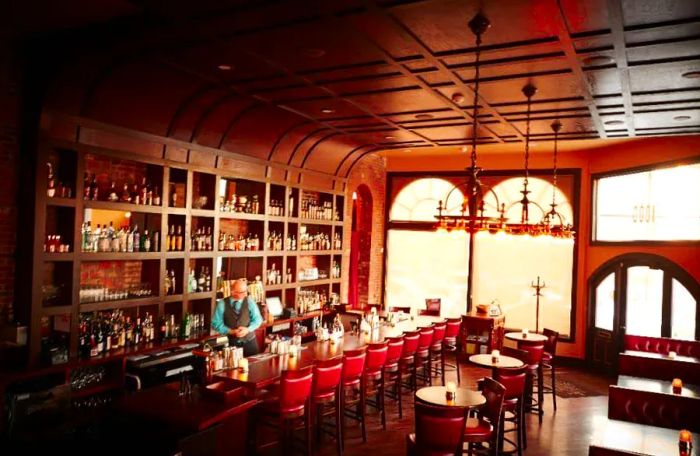 A bartender mixes drinks behind a wooden bar in a cozy, wood-paneled room featuring large windows, red leather stools, and a variety of tables and booths.