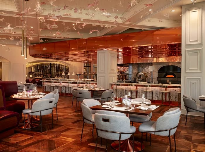 A dining space featuring wide midcentury chairs around wooden tables, accented with copper and marble in the bar area, which boasts an open wood-fired oven against the bar wall, and decorative cranes suspended from the ceiling.