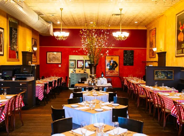 A vibrant dining room filled with four-top tables adorned with checkered tablecloths, featuring a large pot of flowers at the center and decorative posters on the walls.