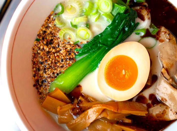 A bowl of ramen featuring a rich, milky broth, a hard-boiled egg, scallions, slices of meat, seeds, bok choy, and burdock root