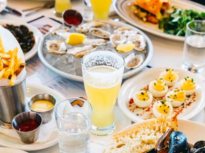 A table overflowing with food and drinks, featuring deviled eggs, oysters on ice, mussels, sandwiches, fries, and hearty steins of beer.