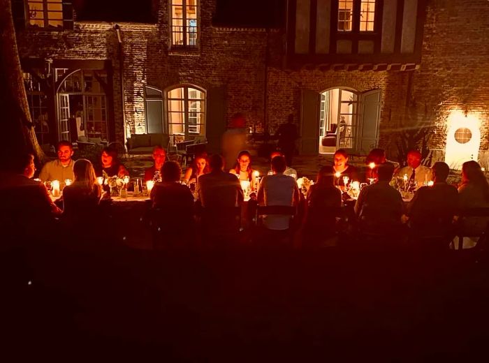 Diners gathered around a long, candlelit table set before an impressive mansion.