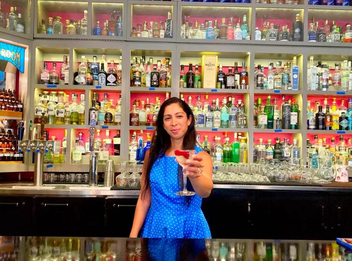 A bartender passes a vibrant cocktail over the bar, with a wide array of illuminated spirit bottles in the background.