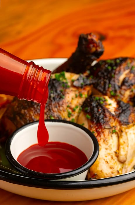 Banana ketchup flows from a bottle into a small ramekin placed next to a roasted chicken.