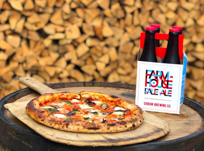 A four-pack of beer bottles placed next to a whole pizza on a wooden paddle, all set on a barrel in front of a stacked wood pile