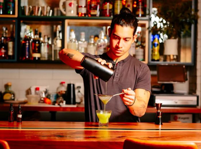 A bartender pours a cocktail from a shaker through a fine mesh strainer into a lowball glass, with a backbar filled with bottles behind.