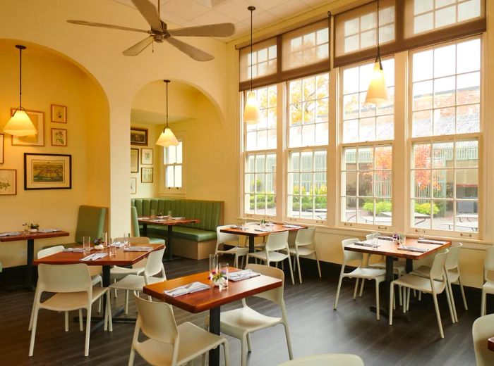 A spacious dining room featuring high ceilings, large windows that bring in ample light, two-top tables set for dinner with casual seating, and arches that separate various dining areas.