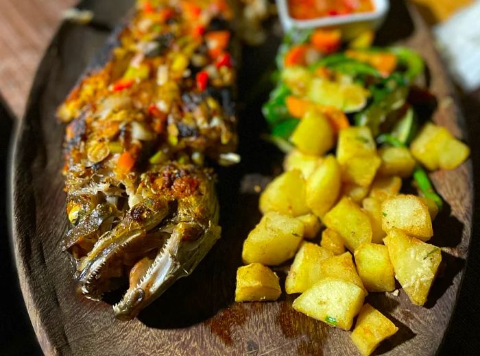 A whole fish served under a hearty sauce alongside crispy potato wedges.