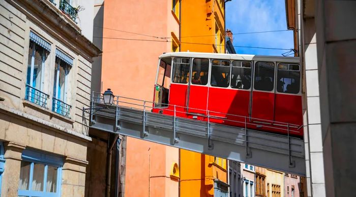 A red funicular train travels along an elevated track between buildings in Lyon.